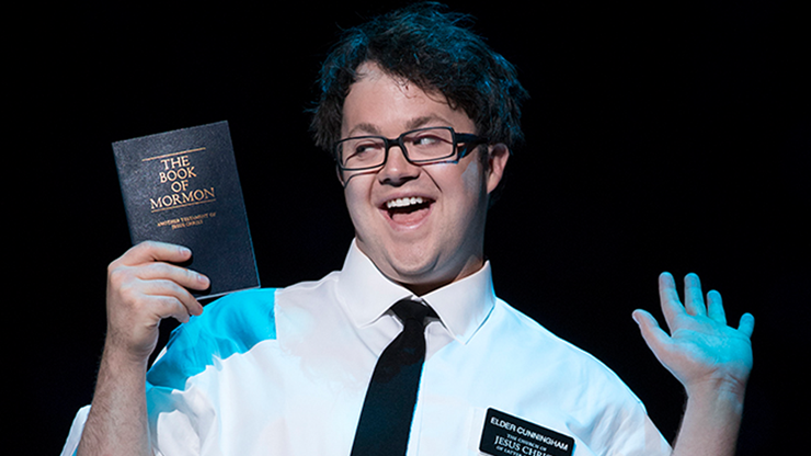 A picture of Cody Jamison Strand with a spotlight on him. He has his hands up and is holding a booklet that says The Book of Mormon. He is wearing a white shirt with a black tie and has a nametag that says Elder Cunningham. 