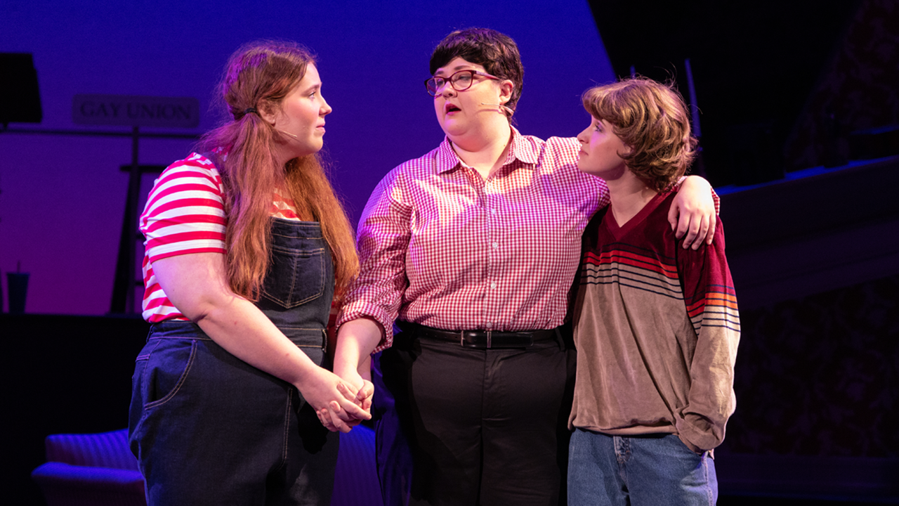 Three theatre students in costume hold their arms around each other and look at each other on stage.
