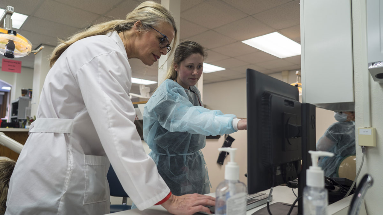 Two women in dental scrubs look and point at a computer screen.