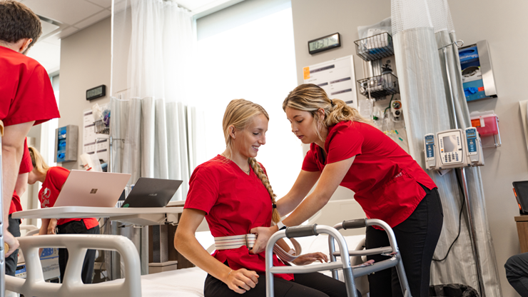A nursing student wraps a strap around another nursing student to help them stand up out of a bed. The nursing students are in a nursing classroom and wear red scrubs.