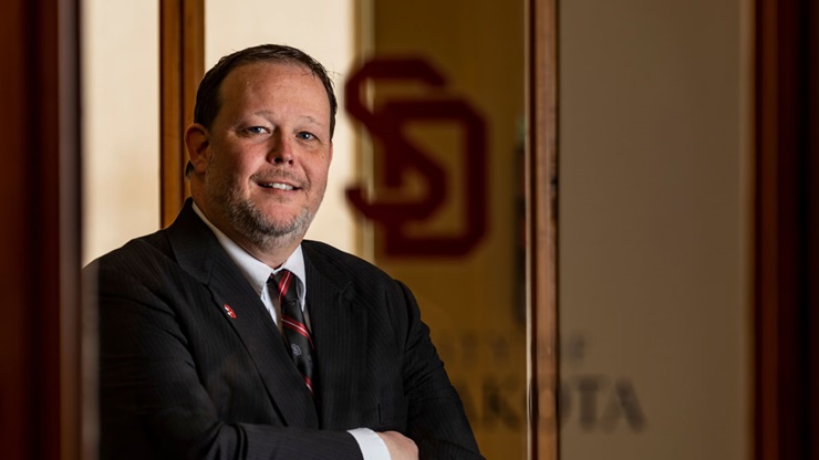 Jay Perry stands in front of a door at USD - Sioux Falls with his arms crossed and he is smiling at the camera. Jay Perry stands in front of a door at USD - Sioux Falls with his arms crossed and he is smiling at the camera.