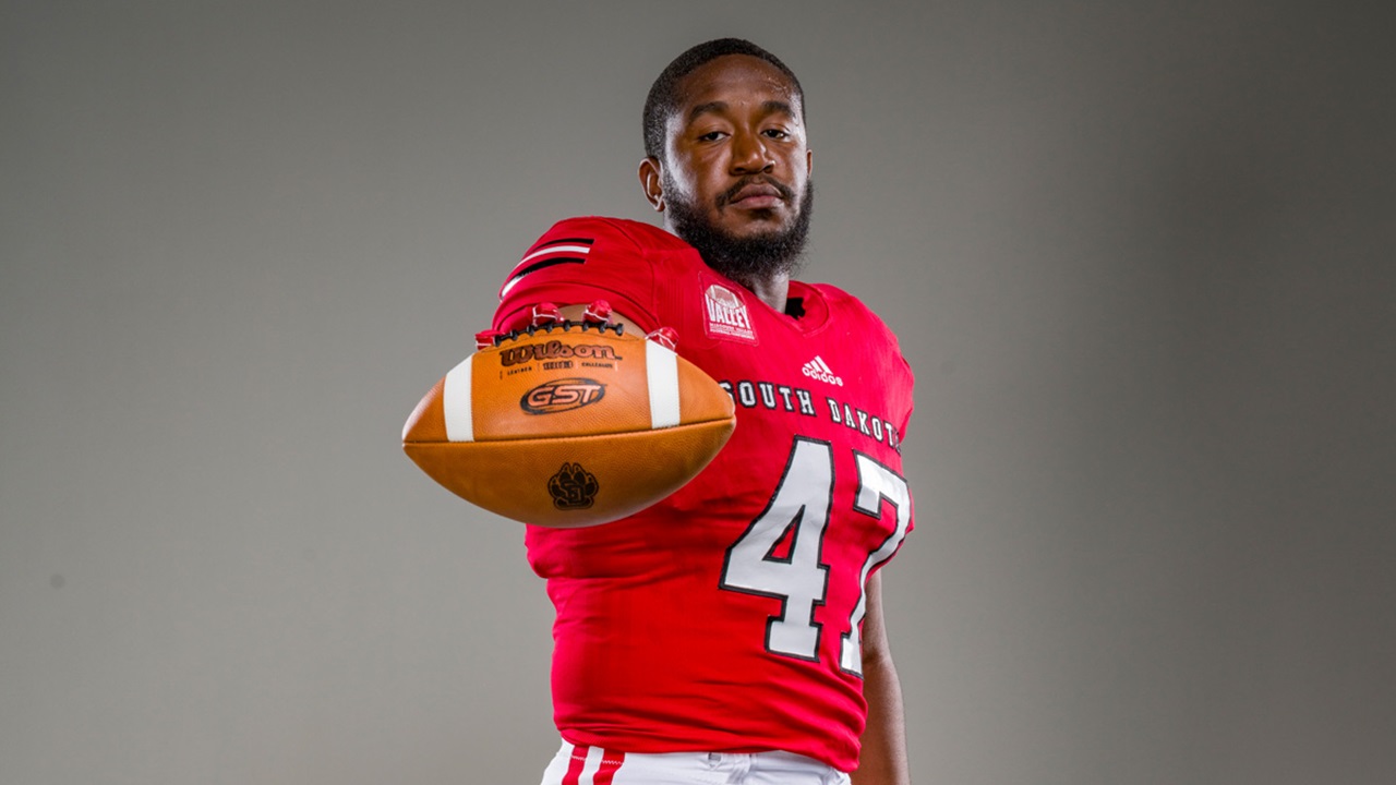 A portrait of DeValon Whitcomb holding a football out in front of him.