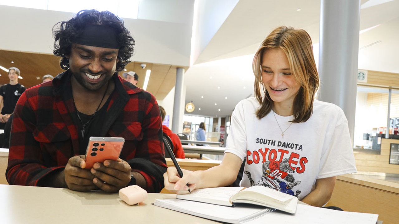 USD students sit at table together. One is working in a notebook. The other is on their phone. USD students sit at table together. One is working in a notebook. The other is on their phone.