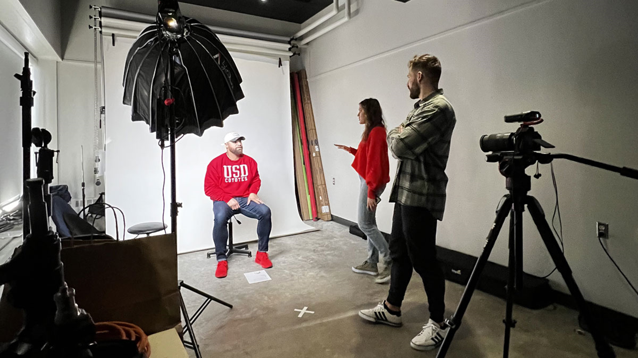 A man sitting on a stool in front of a white background with studio lights and a camera pointing at him. Two people stand to the side and direct him.