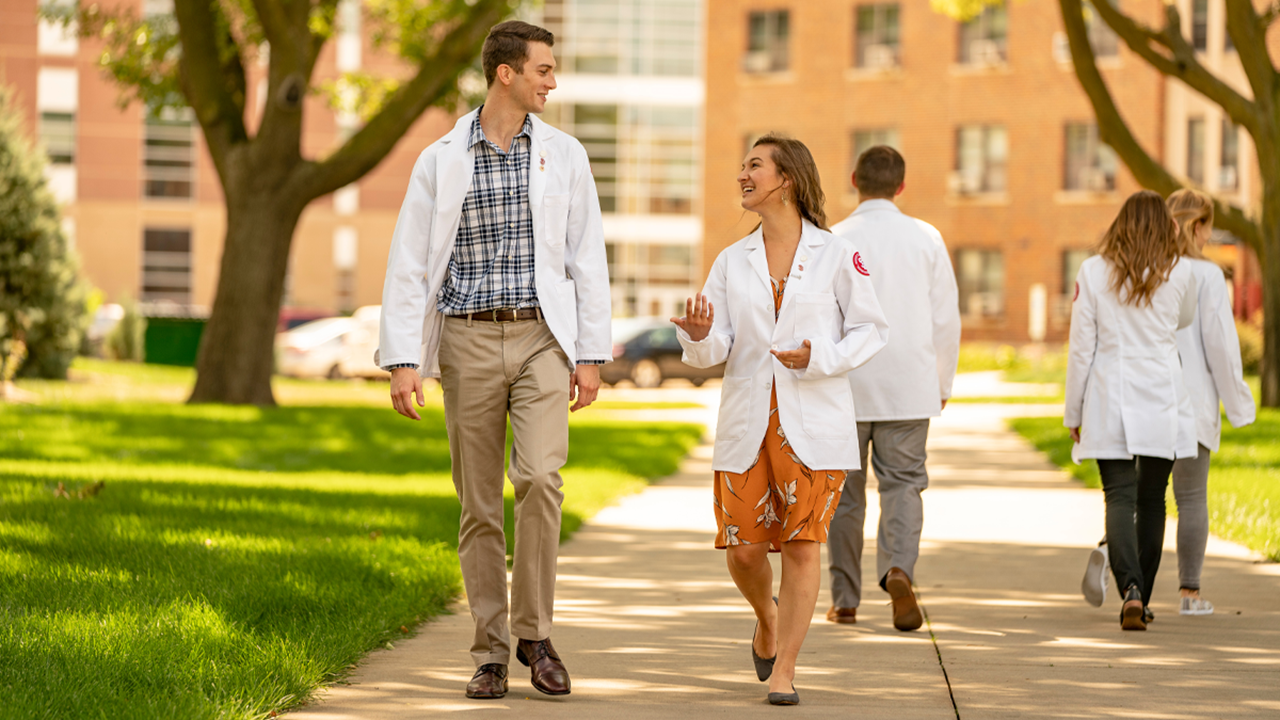Two students walk down the sidewalk wearing white medical coats and talking to each other. Another student in the background behind them walks away from the camera.