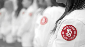 A close up photo of a line of people's shoulders wearing white medical coats. The whole photo is black and white except for the red USD Medical School emblems on the side of the coats.