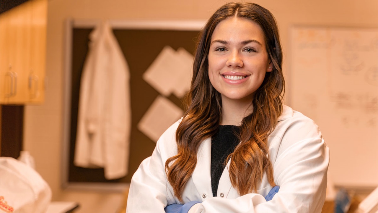 A portrait of Emily Eisenbraun smiling in a lab. She is wearing a white lab coat.