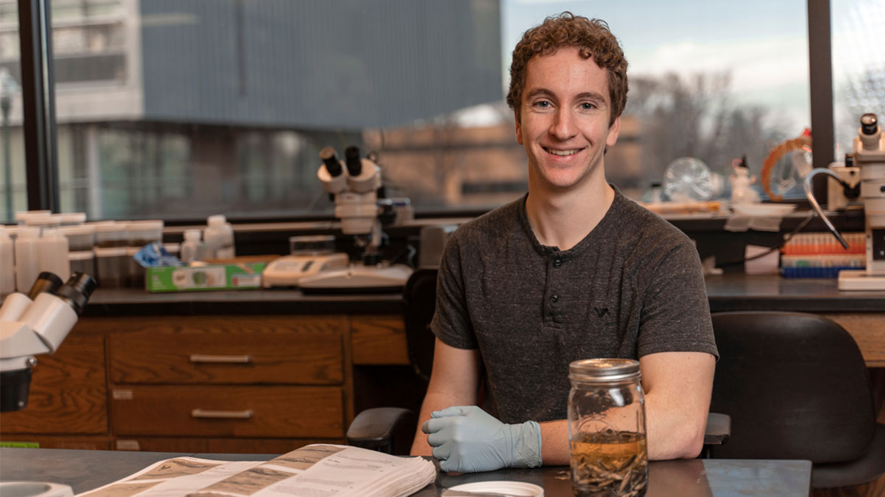Jacob Ridgway sits at a lab desk and smiles. He has a jar with liquid in it and a book in front of him on the table.