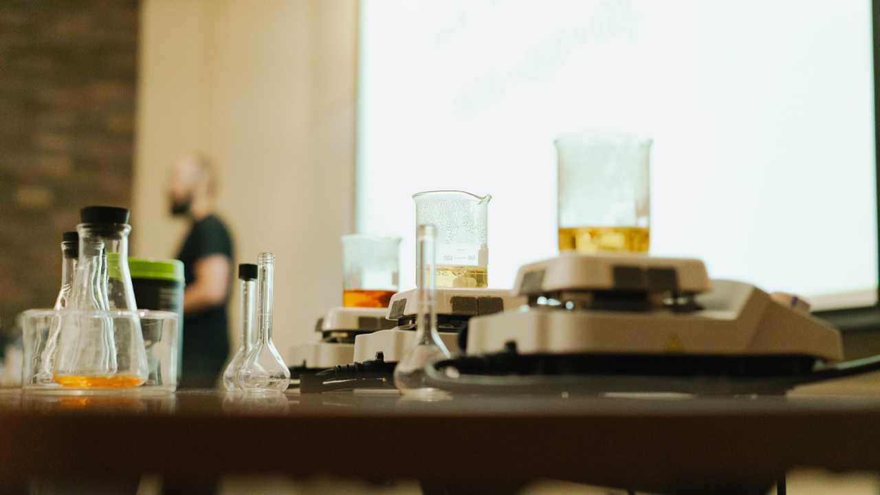 Chemistry beakers and flasks filled with various liquids sit on a desk in a classroom. A professor presents in the background.
