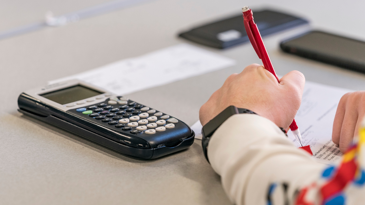 A close-up shot of a student writing on a piece of paper with a red mechanical pencil. There is a calculator sitting to the side of the paper.