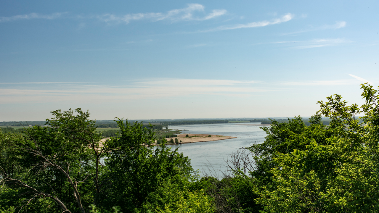 A wide-angle shot of the Missouri River. The sky is blue and the river is lined with green trees.
