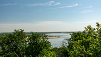 A wide-angle shot of the Missouri River. The sky is blue and the river is lined with green trees. 