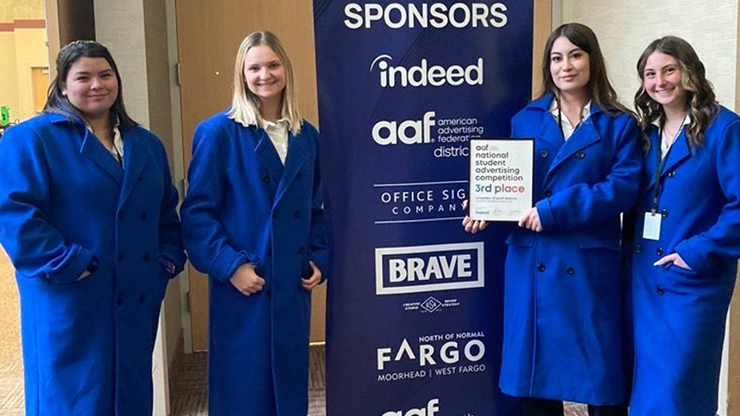 Four female students wear blue robes and pose with a 3rd place plaque from the American Advertising Federation.
