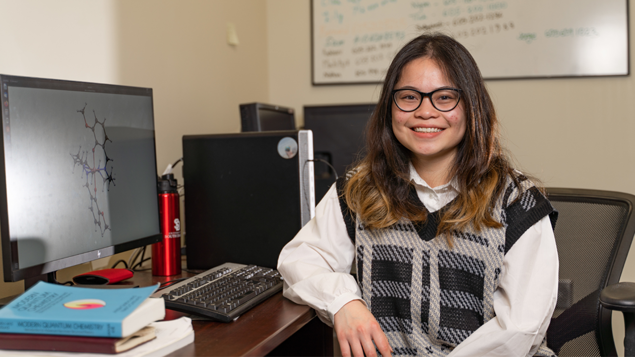 A student sitting at a desk with books and a computer behind them.