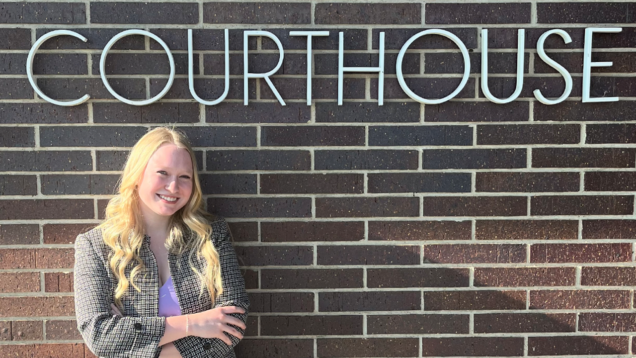 A student stands with their arms crossed in front of a brick wall with the word courthouse written on it.