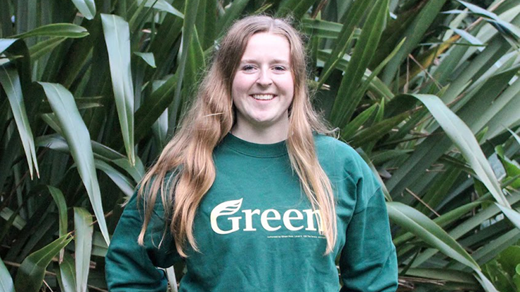 A person wearing a green sweatshirt with the word green across the front stands in front of a corn field and smiles.