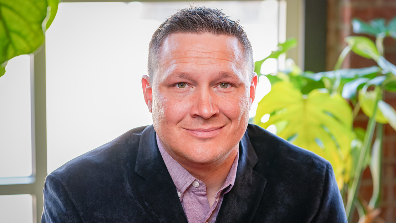 Rocky Welker looks at the camera with a soft smile. He is wearing a suit jacket and sits in front of a window and green plants.