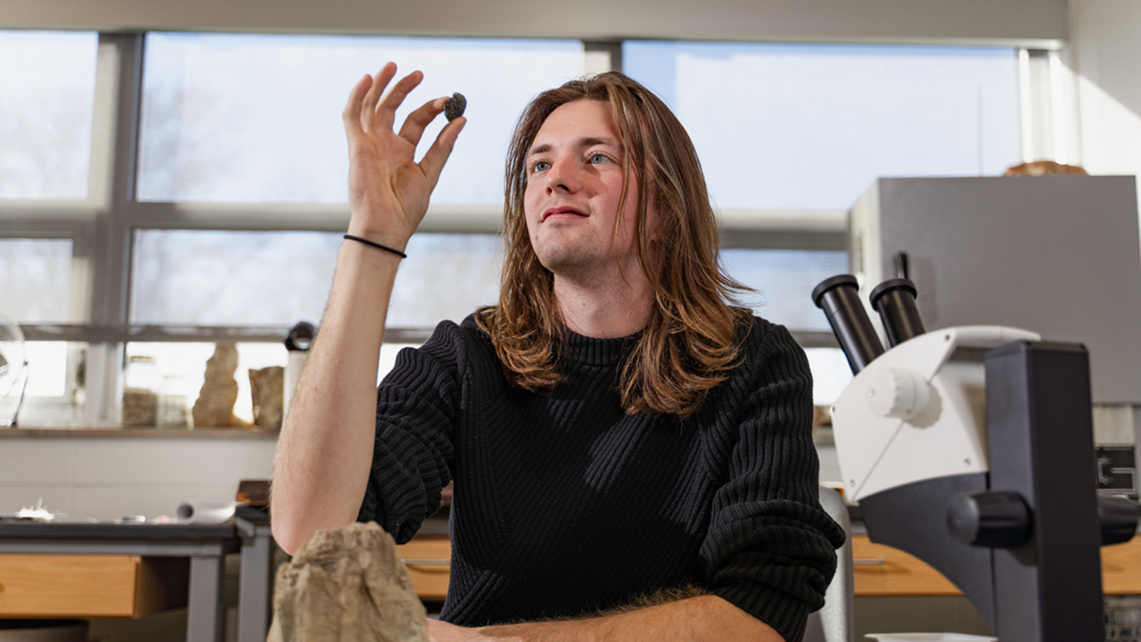 A photo of Wyatt Wiebelhaus holding up a rock and examining it. He is in a lab and there is a microscope in front of him to the right.