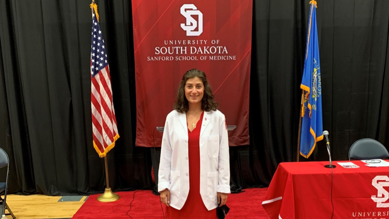 Anna Myrmoe wears her white coat and stands in front of a red University of South Dakota Sanford School of Medicine flag.