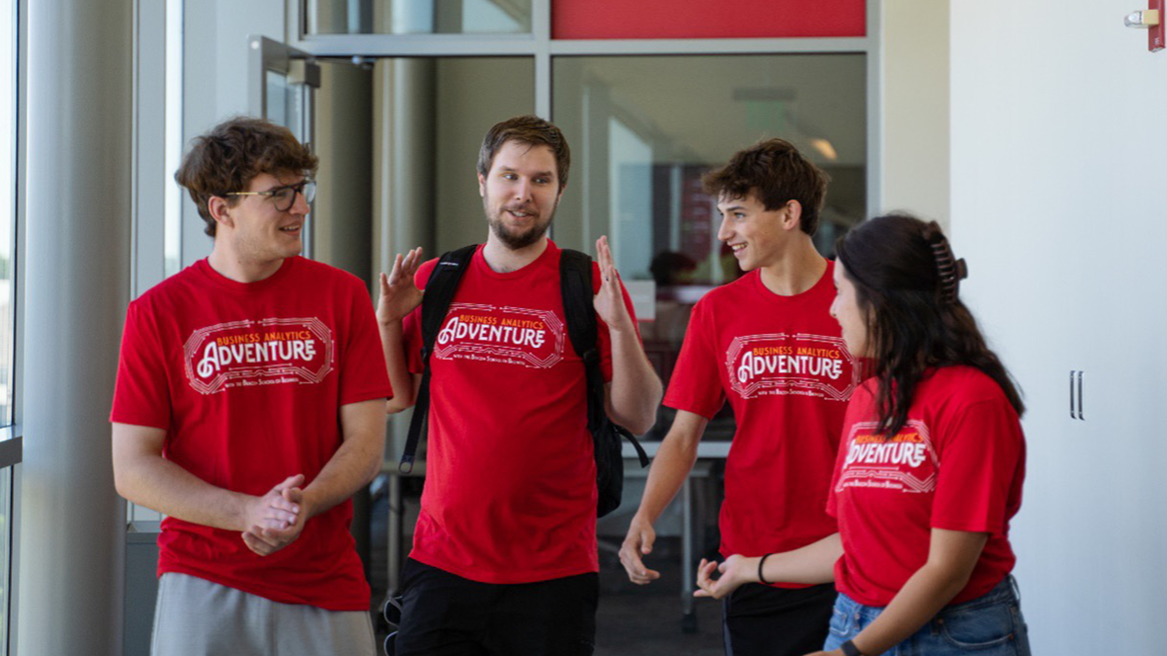 Four students walk together through a hallway and talk. They are all wearing red t-shirts that say Business Analytics Adventure.