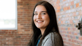 Abigail Boysen looks at the camera and smiles. She is standing in a room with brick walls and a window.