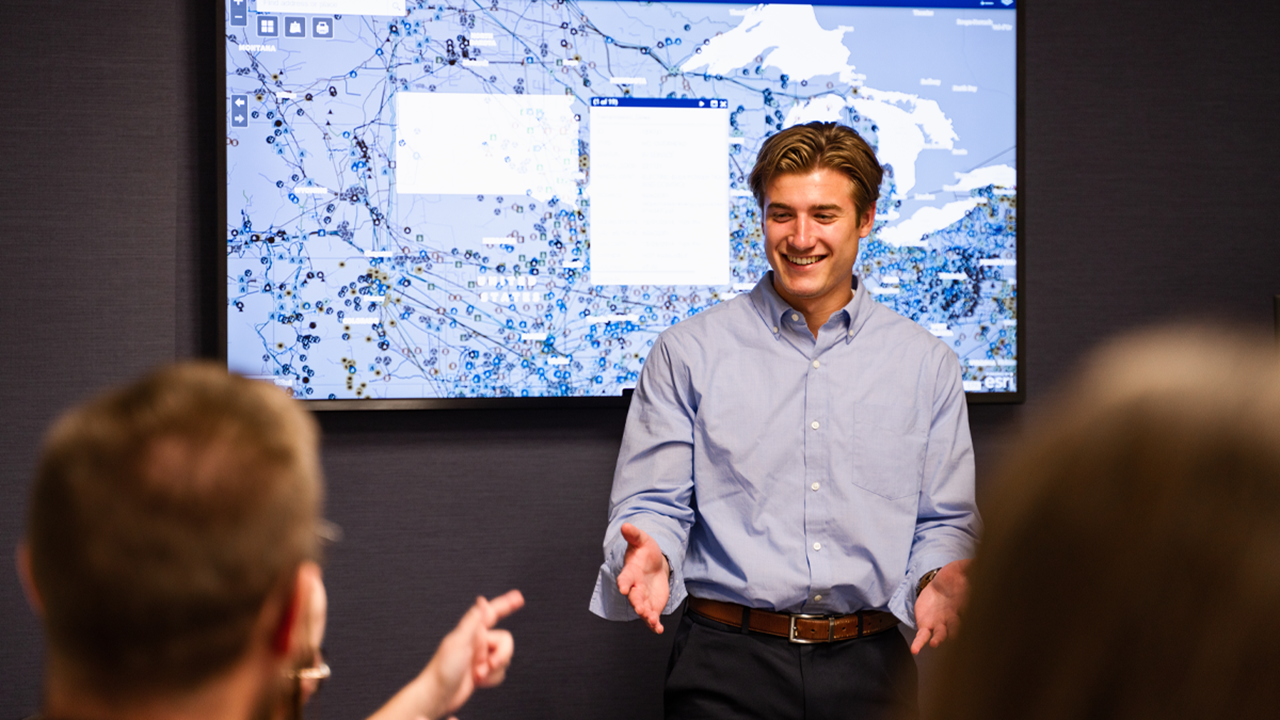 A student stands in front of a screen and presents to a table full of people. The student smiles and holds their hand out to a person sitting at the front of the table.