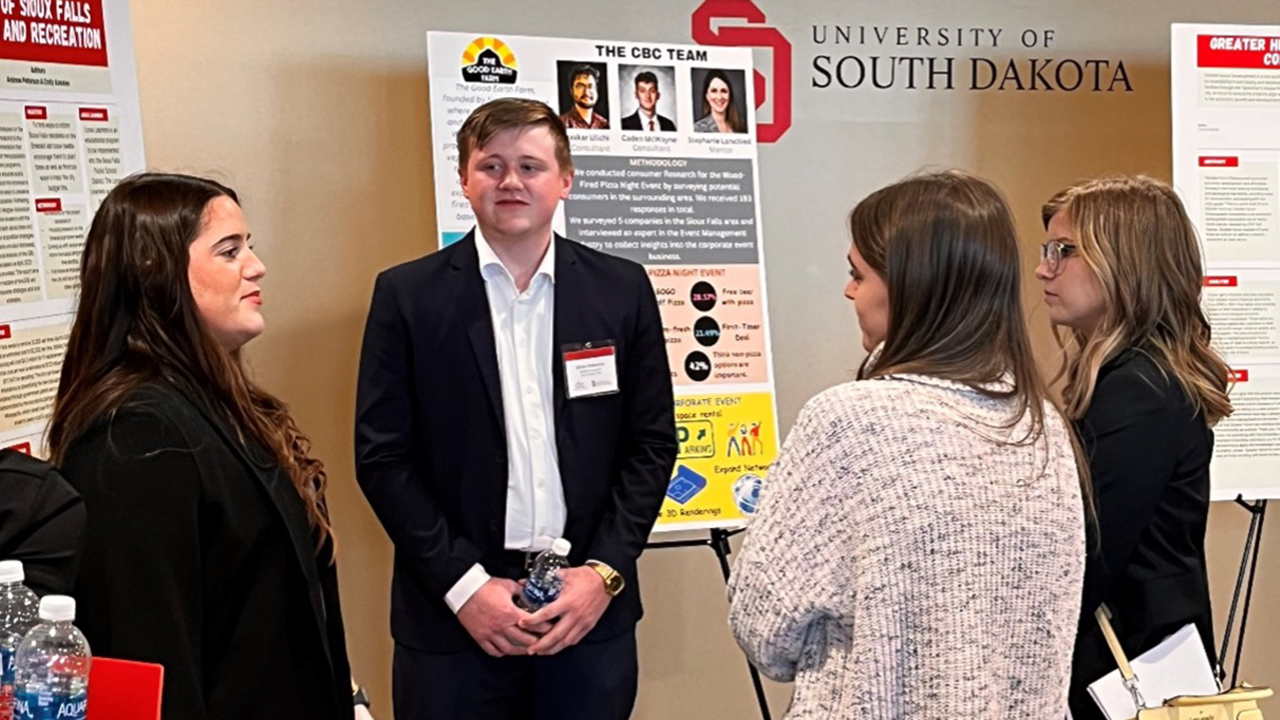 Students wear business professional clothing and stand in front of posters. Two students present information while the other two stand and listen.