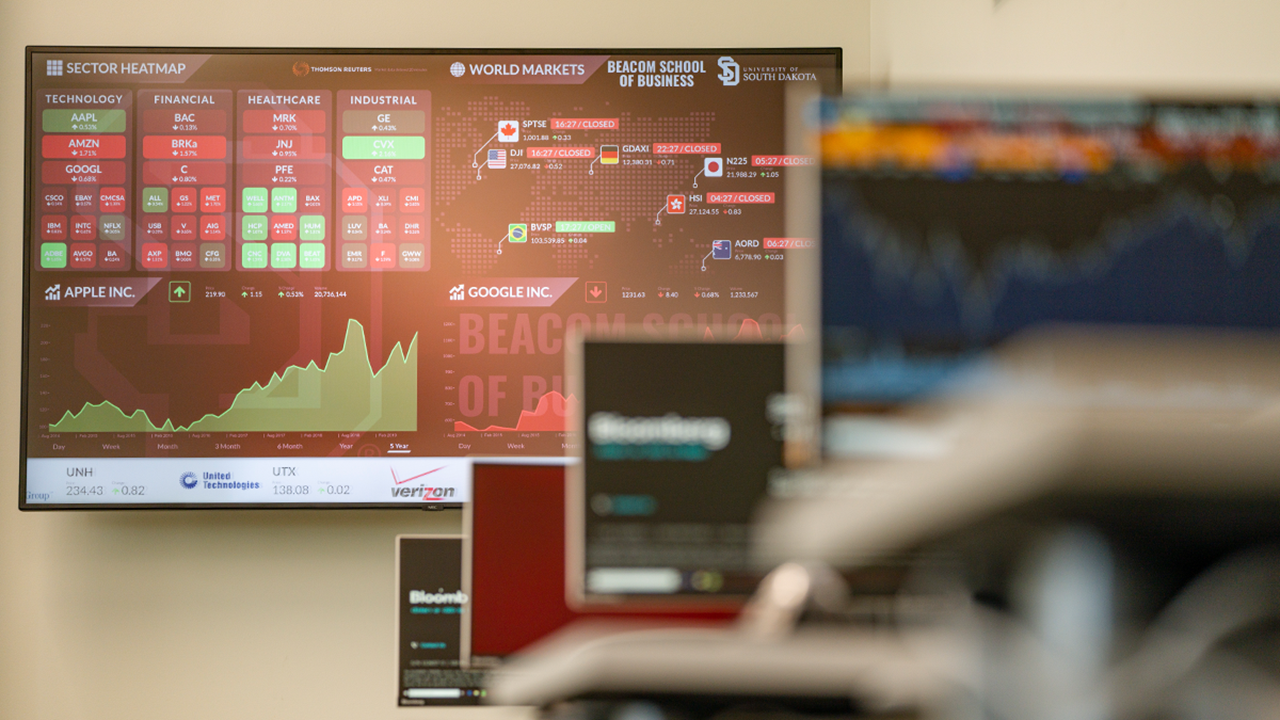 A large TV screen and smaller computer screens show stocks and Bloomberg Terminals.