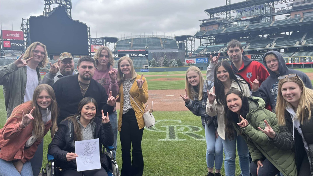 Thirteen college students stand in front of Coors Field in Denver.