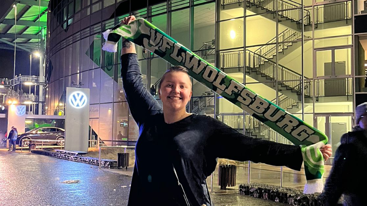 A student holds up a green scarf that says VF Wolfsburg. She stretches her arms out and smiles. She is standing in front of a soccer arena.