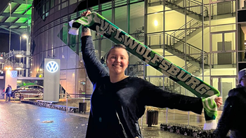 A student holds up a green scarf that says VF Wolfsburg. She stretches her arms out and smiles. She is standing in front of a soccer arena.