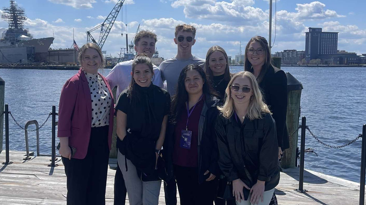 Business students stand together for a photo in front of a large body of water. They stand on a wooden platform and there is a cityscape behind them.