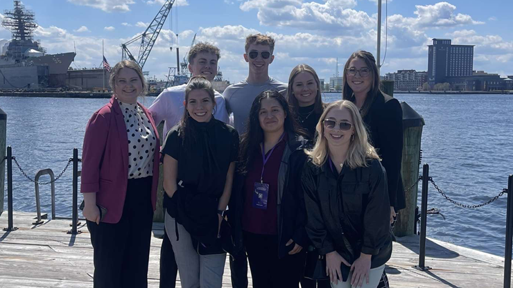 Business students stand together for a photo in front of a large body of water. They stand on a wooden platform and there is a cityscape behind them. 