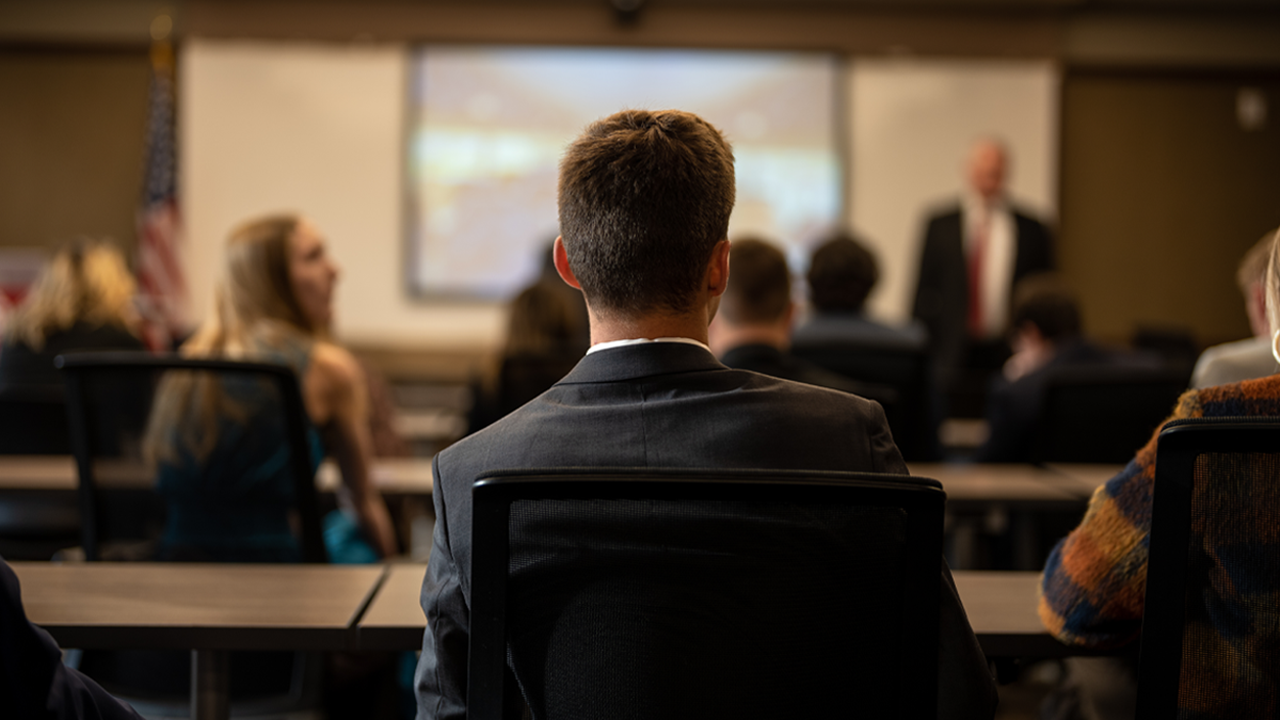The back of a student's head as they look forward in a classroom. There are rows of desks with other students, also facing forward.