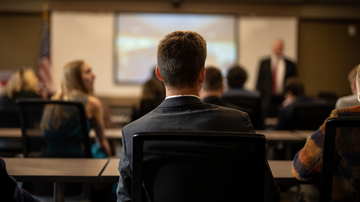 The back of a student's head as they look forward in a classroom. There are rows of desks with other students, also facing forward. 