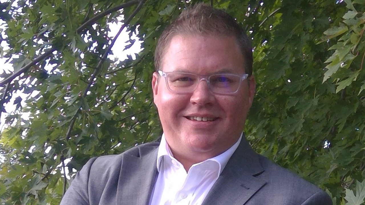 Eric Leise looks forward and smiles. He is standing in front of a tree with green leaves.