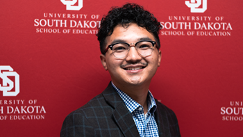A headshot of Hser Lar Kpaw Htoo. He is standing in front of a red background that has white University of South Dakota School of Education text on it.