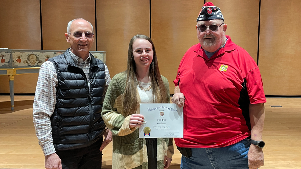 A female student stands between two men who belong to the Veterans of Foreign Wars Organization. The student holds a first place certification.