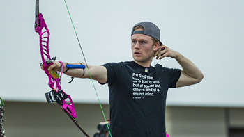 Robby Weissinger stands in an archery stance with one arm bent behind his head and the other straightforward holding a pink bow.