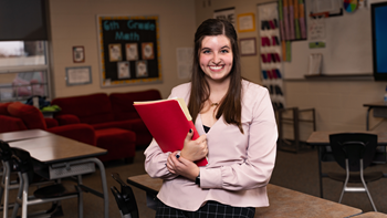 A photo of a student leaning on a desk in a classroom. They hold a red folder in their arms.
