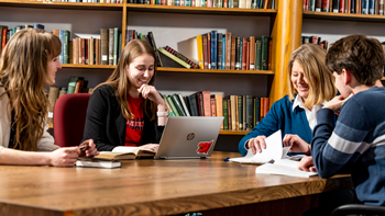 Three English students and one professor sit around a wooden table and talk with one another. There is a bookshelf in the background.