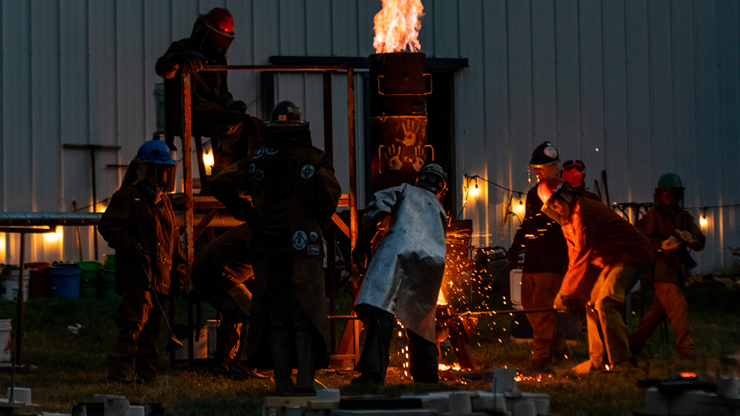 Artists wear heat-protectant clothing and face shields and engage in an iron pour. They stand around a mold and pour molten iron. It is dark outside.