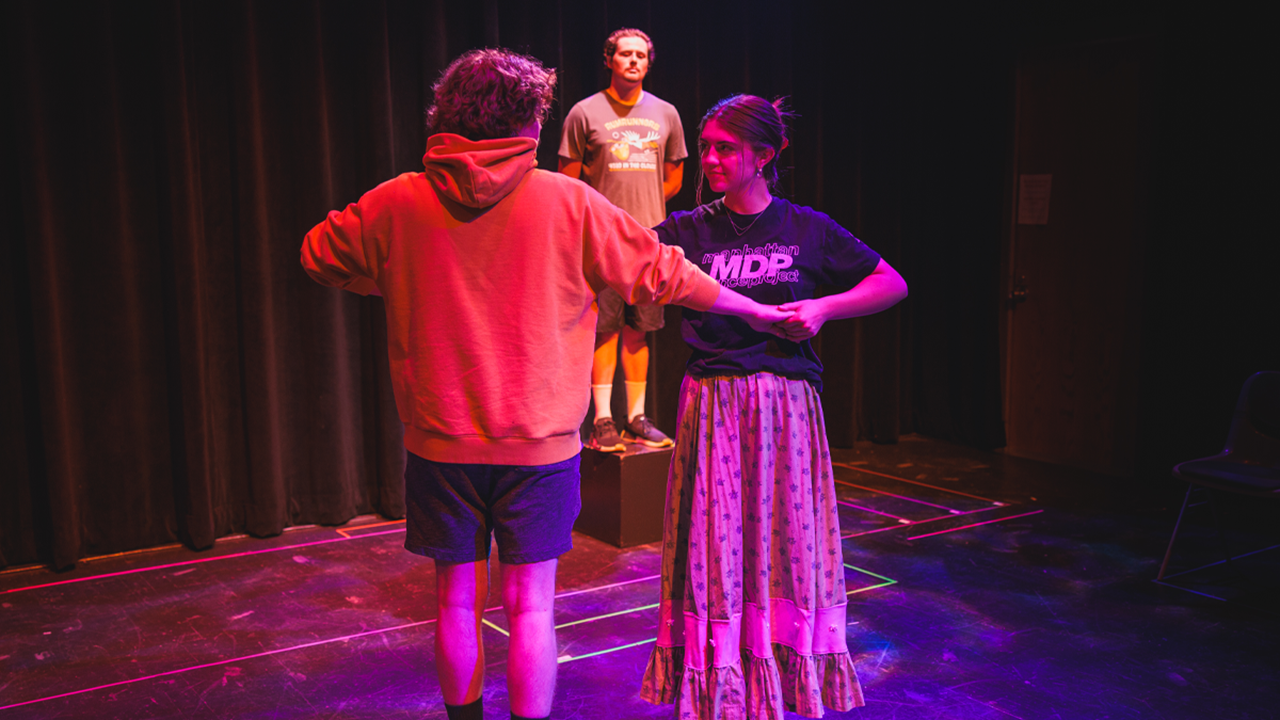 Two students lock hands and dance in a circle on a stage that is lit with colorful lights. A student stands behind and looks at them.