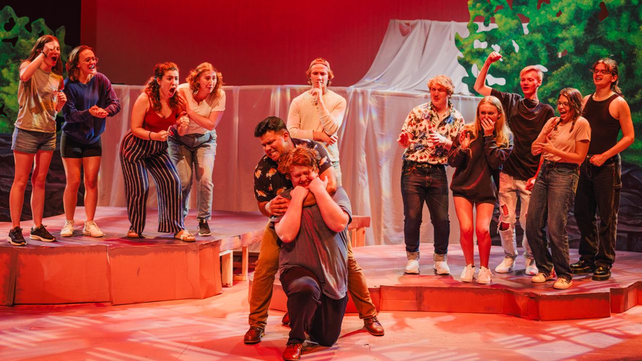 Several actors stand back stage as two actors on front stage get into a physical altercation. One actor has his arm around the other's neck as they work to get free.