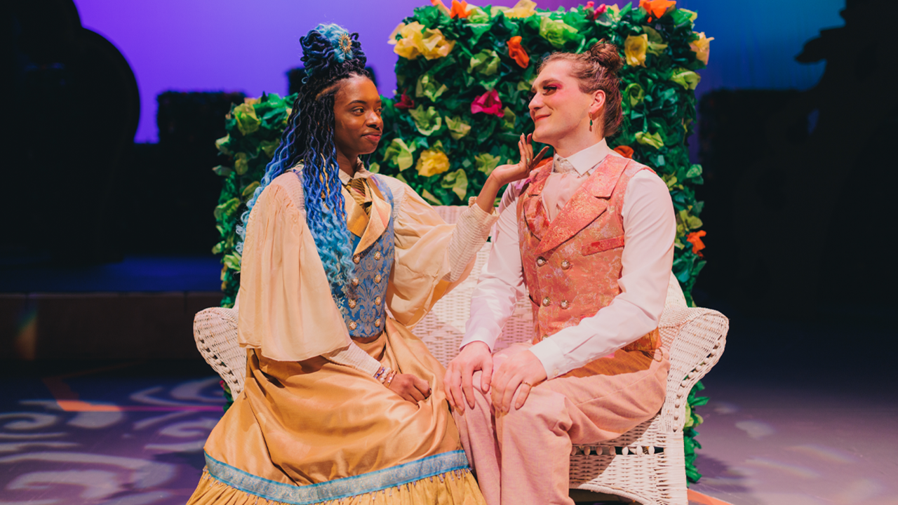 Two theatre students sit on a white whicker bench and face each other. The student on the left is wearing a yellow dress and the student on the right is wearing a pink suit. The background is floral.