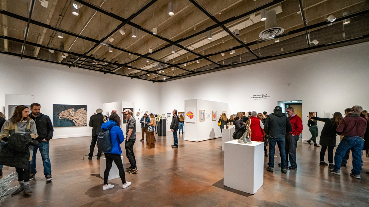 A large group of people stand around and observe art in a gallery.