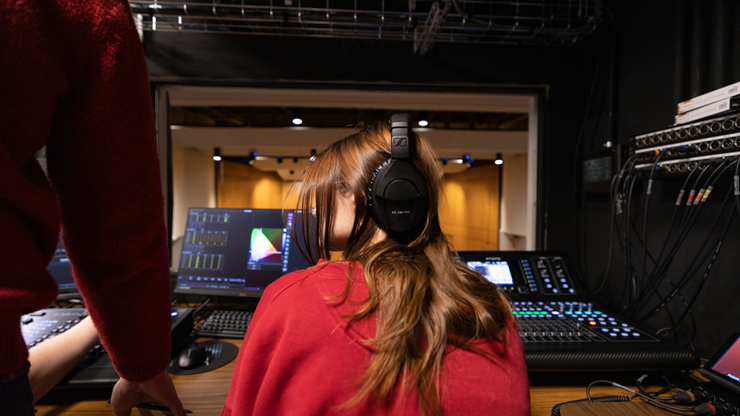 A student wears over-the-ear headphones and sits in front of a music soundboard. A professor's arm leans over to help the student use the board.