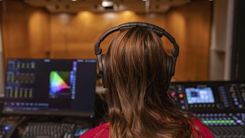 A USD music student wears headphones and is sitting behind a sound board watching and perfecting the sound of a performance.
