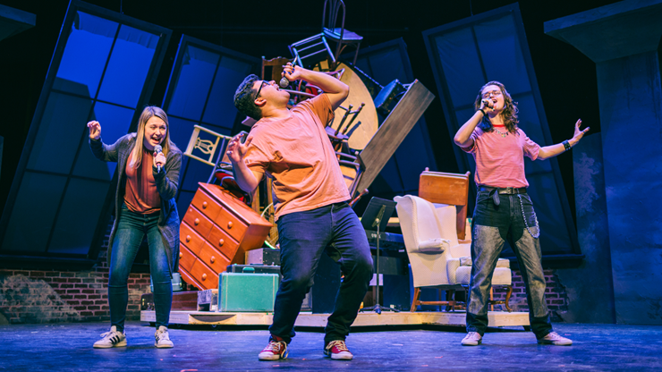 Three musical theater students perform on stage, arms outstretched, surrounded by a stack of props and furniture.
