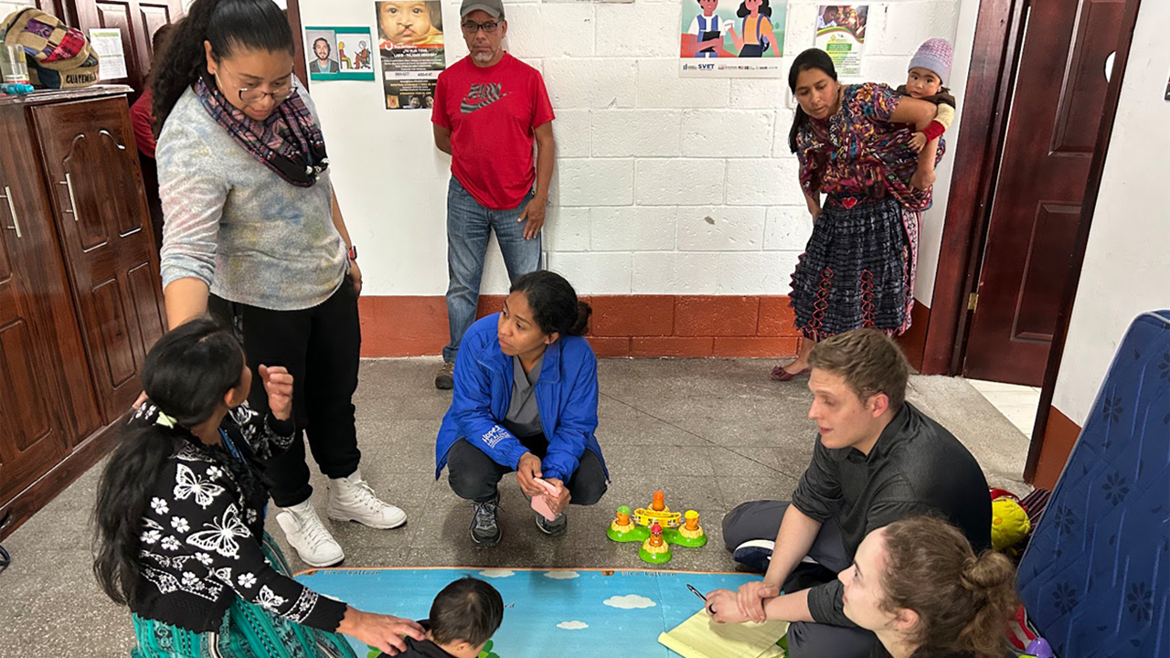 Physical therapy students work with families and children in a classroom setting. Some of them are sitting on the ground learning with toys while other people stand and observe.